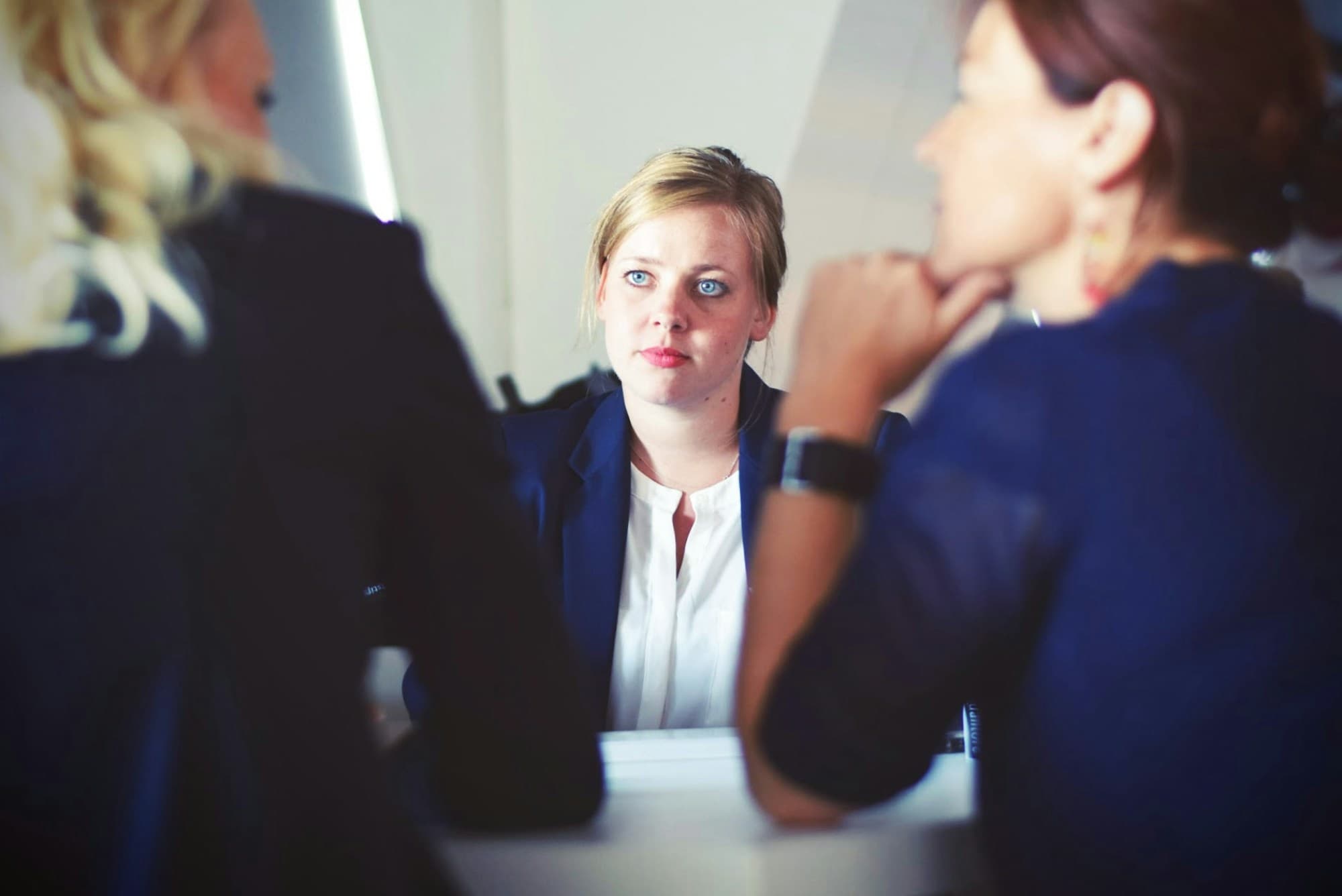 A woman holds a steady, composed expression while responding to two colleagues seated across from her.
